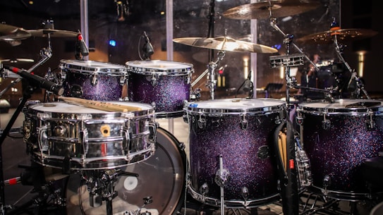 A drum set with sparkling dark purple shells, surrounded by various cymbals and microphones, is featured in a stage setting. The lighting reflects off the shiny surfaces, creating a vibrant and lively atmosphere.