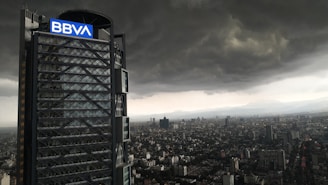A tall modern skyscraper with a prominent 'BBVA' logo is seen next to a sprawling city landscape. The sky is filled with ominous dark clouds, suggesting an impending storm over the urban environment.