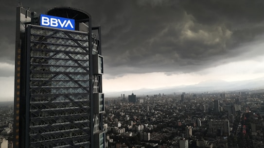 A tall modern skyscraper with a prominent 'BBVA' logo is seen next to a sprawling city landscape. The sky is filled with ominous dark clouds, suggesting an impending storm over the urban environment.
