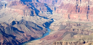 A vast, expansive view of the Grand Canyon with its rugged, layered rock formations and a river winding through the canyon floor. The image showcases the canyon's immense scale and depth, with varying shades of red, brown, and tan rock formations.
