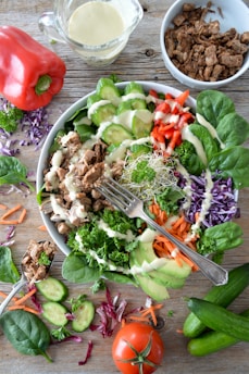 A vibrant and colorful salad arranged in a bowl, featuring a mix of fresh vegetables including sliced cucumbers, avocado, red pepper strips, carrot shreds, and leafy greens topped with creamy dressing. Shredded meat is scattered on top, complemented by garnishes of parsley and sprouts. Nearby, there are additional ingredients like a whole red bell pepper, chopped red cabbage, a ripe tomato, sliced cucumbers, and cooked meat in a separate bowl.
