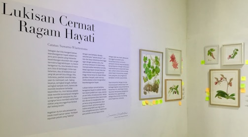 An exhibition wall featuring botanical illustrations with a descriptive text on the left. The text panel includes several paragraphs in Indonesian, and on the right are framed botanical art pieces depicting various plants and flowers. Small colored sticky notes are attached below and beside these frames.