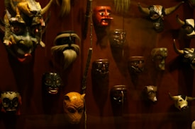 A collection of various masks is displayed on a dark red background. The masks exhibit a range of expressions and designs, including a dragon-like mask with horns, a red-faced mask, and several animalistic and human-like masks. Some masks feature intricate patterns, while others have more simplistic designs.