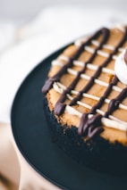 A close-up view of a chocolate-covered cheesecake with a crumbly black crust. The surface is decorated with a lattice of dark and white chocolate drizzles. The cake is placed on a dark plate, with a soft focus on the background.