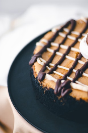 A close-up view of a chocolate-covered cheesecake with a crumbly black crust. The surface is decorated with a lattice of dark and white chocolate drizzles. The cake is placed on a dark plate, with a soft focus on the background.