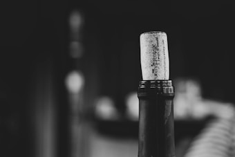 A close-up of a wine bottle with a cork partially inserted into its neck. The background is blurred, keeping the focus on the bottle and cork, creating a sense of depth. The image is in black and white, giving it a classic and timeless feel.