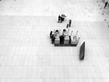 A large, open, tiled floor space is centered upon a small cluster of people engaged with several stacked trolleys containing various items. There's a sense of organization and preparation, as equipment or materials are being handled. The perspective is from above, offering a bird’s-eye view.