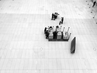 A large, open, tiled floor space is centered upon a small cluster of people engaged with several stacked trolleys containing various items. There's a sense of organization and preparation, as equipment or materials are being handled. The perspective is from above, offering a bird’s-eye view.