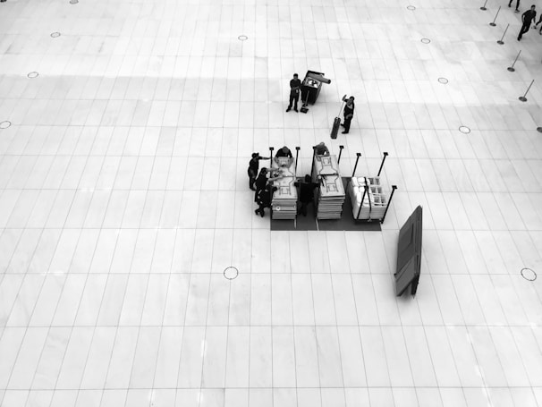 A large, open, tiled floor space is centered upon a small cluster of people engaged with several stacked trolleys containing various items. There's a sense of organization and preparation, as equipment or materials are being handled. The perspective is from above, offering a bird’s-eye view.