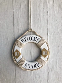 A decorative life ring hangs against a textured wall. The ring is adorned with rope and features the text 'Welcome Aboard' in blue letters, with two yellow diamond shapes on either side containing the number '01'.