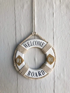 A decorative life ring hangs against a textured wall. The ring is adorned with rope and features the text 'Welcome Aboard' in blue letters, with two yellow diamond shapes on either side containing the number '01'.