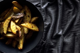 A dark bowl filled with seasoned potato wedges and bay leaves sits on a textured, dark fabric background. The lighting emphasizes the golden, crispy texture of the potatoes and the dark, moody setting creates a rustic and savory atmosphere.
