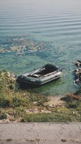 A small inflatable boat is resting on the edge of a lake with clear green-blue water. The shoreline is lined with grass and rocks, and there are some water plants visible near the boat. The ripples in the water create a peaceful and serene atmosphere.