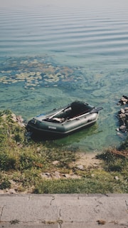 A small inflatable boat is resting on the edge of a lake with clear green-blue water. The shoreline is lined with grass and rocks, and there are some water plants visible near the boat. The ripples in the water create a peaceful and serene atmosphere.