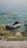 A small inflatable boat is resting on the edge of a lake with clear green-blue water. The shoreline is lined with grass and rocks, and there are some water plants visible near the boat. The ripples in the water create a peaceful and serene atmosphere.