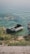 A small inflatable boat is resting on the edge of a lake with clear green-blue water. The shoreline is lined with grass and rocks, and there are some water plants visible near the boat. The ripples in the water create a peaceful and serene atmosphere.
