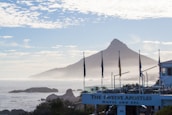 A scenic view of a coastal landscape with a prominent mountain in the background. Flags are lined up in front of a building that has 'The Twelve Apostles Hotel and Spa' sign. The ocean is visible with rocks and mist adding to the serene atmosphere. The sky is partly cloudy, creating a peaceful, tranquil setting.