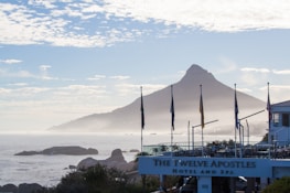A scenic view of a coastal landscape with a prominent mountain in the background. Flags are lined up in front of a building that has 'The Twelve Apostles Hotel and Spa' sign. The ocean is visible with rocks and mist adding to the serene atmosphere. The sky is partly cloudy, creating a peaceful, tranquil setting.