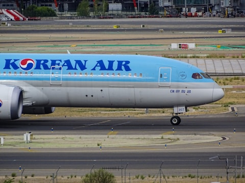 A large passenger airplane with a light blue and white color scheme is taxiing on an airport tarmac. The plane has the name 'Korean Air' prominently displayed along the fuselage. The background shows part of the airport with various structures and equipment.