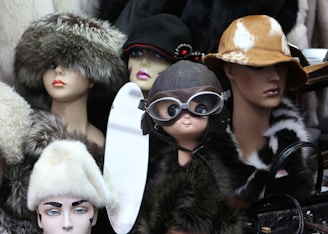 A collection of mannequin heads adorned with various hats and accessories. The hats are made from materials such as fur and leather, ranging from aviator goggles to wide-brimmed styles. Each mannequin exudes a unique character due to the distinct headwear choices, with some displaying elegant flower details and others a more vintage pilot look.