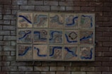 A collection of ceramic tiles mounted on a brick wall, each featuring intricate illustrations with recurring blue river motifs. The tiles depict various scenes and objects, such as birds, landscapes, and abstract designs, with natural and earthy tones emphasized by blue lines.