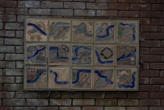 A collection of ceramic tiles mounted on a brick wall, each featuring intricate illustrations with recurring blue river motifs. The tiles depict various scenes and objects, such as birds, landscapes, and abstract designs, with natural and earthy tones emphasized by blue lines.