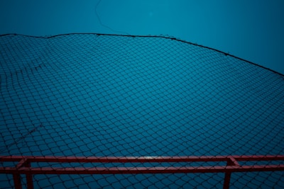 A black net stretches across the frame in a diagonal pattern against a bright blue background. The bottom edge features a red railing or border that provides contrast to the predominant blue. The net is finely woven and creates a geometric, crisscross pattern.