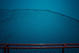A black net stretches across the frame in a diagonal pattern against a bright blue background. The bottom edge features a red railing or border that provides contrast to the predominant blue. The net is finely woven and creates a geometric, crisscross pattern.