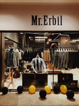 A fashionable store display featuring suits and menswear. Two mannequins hold classic suits with ties, surrounded by a range of neatly arranged suits on racks. Large letters spelling 'Mr. Erbil' are prominently displayed at the top of the shop. Black and yellow balloons are arranged on the floor, adding a touch of color to the sophisticated setting.