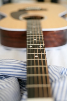A close-up view of an acoustic guitar focusing on the neck and fretboard. The background is softly blurred, highlighting the strings and wood grain texture. The lower portion of the image features blue and white striped fabric, possibly a shirt or bedding.