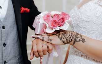 A close-up of a couple's hands, where the woman is wearing a white lace dress with intricate henna designs on her hand and holding a bouquet of pink roses. The man is wearing a black suit with a gray waistcoat and a red pocket square.