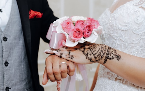 A close-up of a couple's hands, where the woman is wearing a white lace dress with intricate henna designs on her hand and holding a bouquet of pink roses. The man is wearing a black suit with a gray waistcoat and a red pocket square.