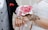 A close-up of a couple's hands, where the woman is wearing a white lace dress with intricate henna designs on her hand and holding a bouquet of pink roses. The man is wearing a black suit with a gray waistcoat and a red pocket square.