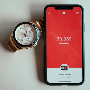A gold and silver wristwatch with a white face and multiple dials lies beside a smartphone. The phone screen displays a red background with the text '70.059 subscribers' and 'Tommy Hilfiger' below it, along with a circular graphic and subscriber view count.