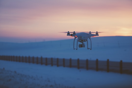 A drone hovers in the air against a scenic backdrop of a sunset sky. The horizon shows a soft gradient of orange, pink, and blue tones as the sun sets over a snow-covered landscape. The ground is flat, and a long fence stretches across the scene, adding depth and perspective to the image.