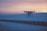 A drone hovers in the air against a scenic backdrop of a sunset sky. The horizon shows a soft gradient of orange, pink, and blue tones as the sun sets over a snow-covered landscape. The ground is flat, and a long fence stretches across the scene, adding depth and perspective to the image.