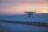 A drone hovers in the air against a scenic backdrop of a sunset sky. The horizon shows a soft gradient of orange, pink, and blue tones as the sun sets over a snow-covered landscape. The ground is flat, and a long fence stretches across the scene, adding depth and perspective to the image.