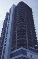 A high-rise building under construction, featuring a modern architectural design with visible concrete structural elements and partially completed exterior. Scaffolding and construction materials are present, indicating ongoing work.