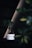 A small bonsai tree with lush green leaves sits in a sleek white ceramic pot. The background features a dark, minimalist setting with subtle diagonal lines and soft lighting. A portion of another plant or leafy structure is visible, adding depth and dimension to the composition.