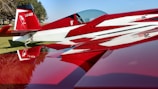 A sleek red and white aerobatic airplane is parked on a grass field. The design features sharp angles and a glossy finish, with a cockpit canopy and distinctive graphics on the tail. Trees and a clear blue sky are visible in the background, creating a picturesque setting.