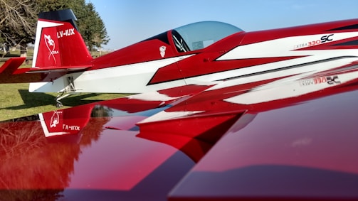 A sleek red and white aerobatic airplane is parked on a grass field. The design features sharp angles and a glossy finish, with a cockpit canopy and distinctive graphics on the tail. Trees and a clear blue sky are visible in the background, creating a picturesque setting.