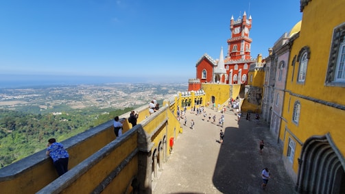 A vibrant and colorful hilltop palace with architectural features painted in bright red and yellow. People are exploring and enjoying the expansive views of the surrounding landscape and sky in the background.
