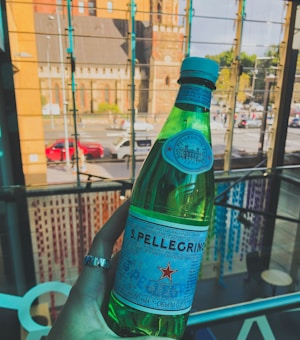 A hand wearing a ring is holding a green bottle of S.Pellegrino sparkling water indoors. In the background, a glass wall reveals an urban street scene with red and white cars, trees, and a tall building. The lighting and reflections create an interesting play of colors and patterns.