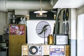 A small retro-themed room with vintage decor featuring vinyl records displayed on the wall, an old-school fan, and a wooden counter with a vinyl disc player. There are drink posters displayed on the wood panel and an amber glass bottle on the counter.