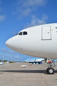 The nose and cockpit window of a large commercial airplane are prominently visible on an airport tarmac under a clear blue sky. A smaller aircraft with the TUI logo is parked in the background along with several airport vehicles and equipment scattered around the scene.