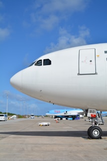 The nose and cockpit window of a large commercial airplane are prominently visible on an airport tarmac under a clear blue sky. A smaller aircraft with the TUI logo is parked in the background along with several airport vehicles and equipment scattered around the scene.