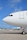 The nose and cockpit window of a large commercial airplane are prominently visible on an airport tarmac under a clear blue sky. A smaller aircraft with the TUI logo is parked in the background along with several airport vehicles and equipment scattered around the scene.