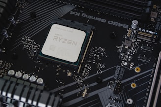 A computer motherboard with a prominently visible AMD Ryzen processor installed. Circuits, transistors, and various electronic components are integrated into the board. The background features a dark, matte texture with intricate patterns and electronic paths.