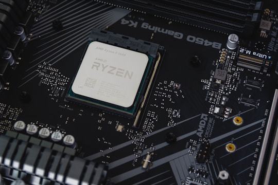 A computer motherboard with a prominently visible AMD Ryzen processor installed. Circuits, transistors, and various electronic components are integrated into the board. The background features a dark, matte texture with intricate patterns and electronic paths.