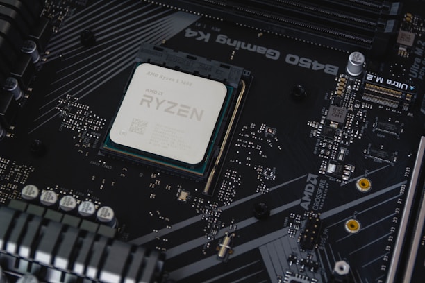 A computer motherboard with a prominently visible AMD Ryzen processor installed. Circuits, transistors, and various electronic components are integrated into the board. The background features a dark, matte texture with intricate patterns and electronic paths.